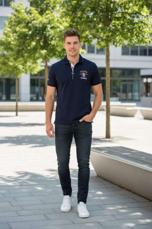 A man wearing a navy blue polo shirt with a white embroidered logo on the left chest, paired with dark gray jeans and white sneakers, 