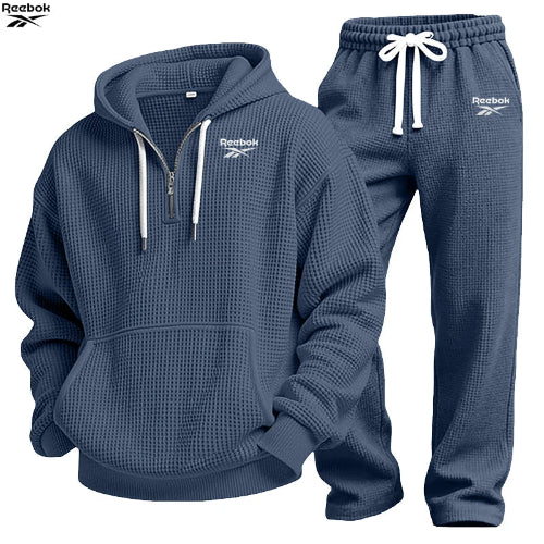 A two-piece Under Armour tracksuit set in navy blue waffle fabric, featuring a half-zip hoodie with white drawstrings and logo details, 