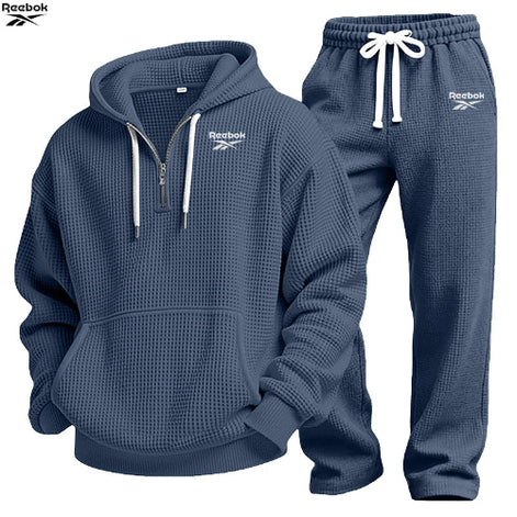 A two-piece Under Armour tracksuit set in navy blue waffle fabric, featuring a half-zip hoodie with white drawstrings and logo details, 