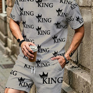 A man wearing a matching light gray t-shirt and shorts set with a black 'KING' crown logo pattern, standing against a stone wall and holding a coffee cup.