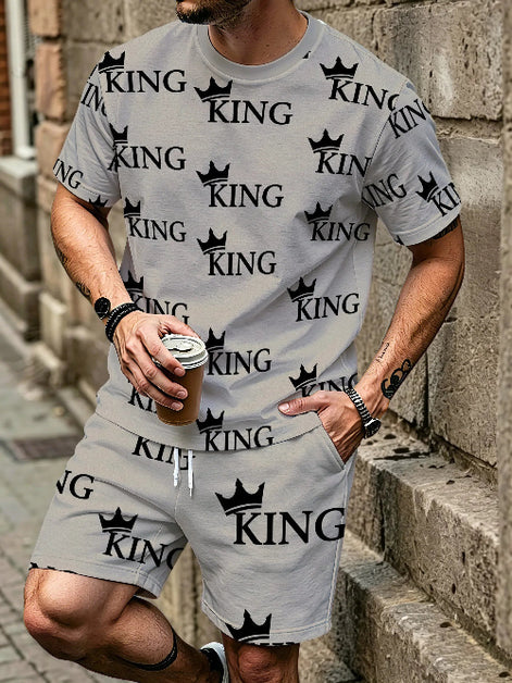 A man wearing a matching light gray t-shirt and shorts set with a black 'KING' crown logo pattern, standing against a stone wall and holding a coffee cup.