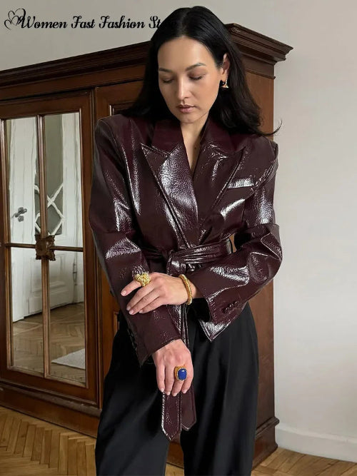 Woman wearing a glossy dark brown belted jacket with wide lapels and buttoned cuffs, adjusting a sleeve beside a wooden cabinet