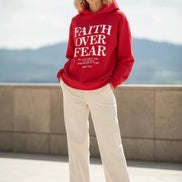 Red hoodie with 'Faith Over Fear' text on a wooden surface with shoes and headphones.