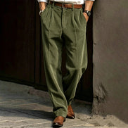 Person wearing green pleated pants and brown shoes against a textured wall.