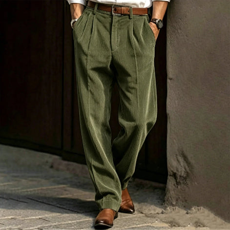 Person wearing green pleated pants and brown shoes against a textured wall.