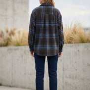A long-sleeve plaid flannel shirt jacket in dark brown, blue, and black, hanging on a hanger on a clothing rack in a boutique.