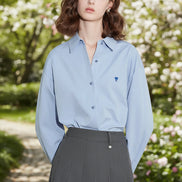 Woman wearing a light blue shirt with a small logo, standing against a white Wall