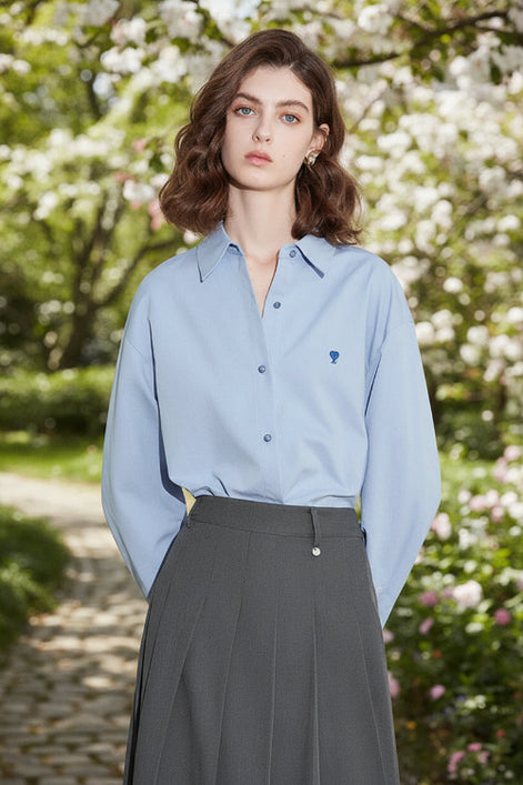 Woman wearing a light blue shirt with a small logo, standing against a white Wall