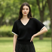 A woman wearing a black short-sleeve V-neck knit top with a white collar and white sleeve trim, paired with a white collared shirt underneath, holding a smartphone and carrying a black shoulder bag.