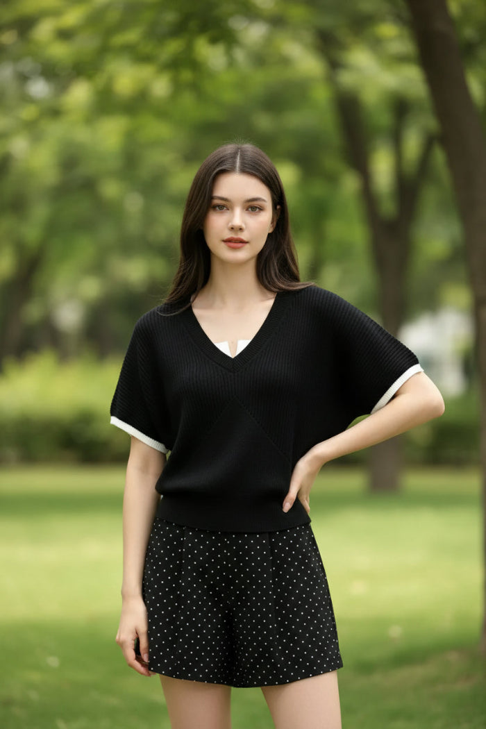 A woman wearing a black short-sleeve V-neck knit top with a white collar and white sleeve trim, paired with a white collared shirt underneath, holding a smartphone and carrying a black shoulder bag.
