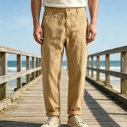 A person standing on a wooden boardwalk by the ocean wearing tan straight-leg trousers, a white t-shirt, and off-white sneakers.