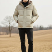 A man wearing a light beige hooded puffer jacket with a brown inner hood lining over a black turtleneck. Three feature callout graphics on the left highlight down insulation, warm thickness, and soft fluffiness.