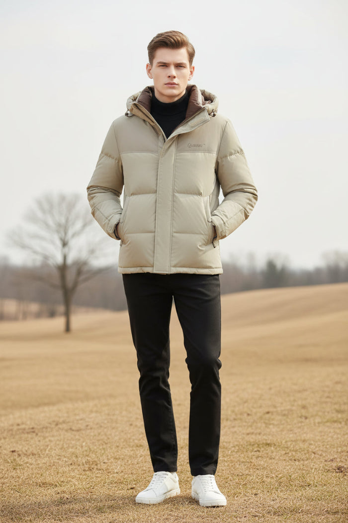 A man wearing a light beige hooded puffer jacket with a brown inner hood lining over a black turtleneck. Three feature callout graphics on the left highlight down insulation, warm thickness, and soft fluffiness.