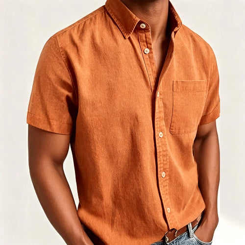A man wearing a short-sleeve button-up shirt in a warm rust orange color, paired with blue jeans against a plain light background.
