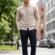 A light beige knit cardigan sweater with a crew neck and button closure, hanging on a dark wooden hanger against a solid gray background.