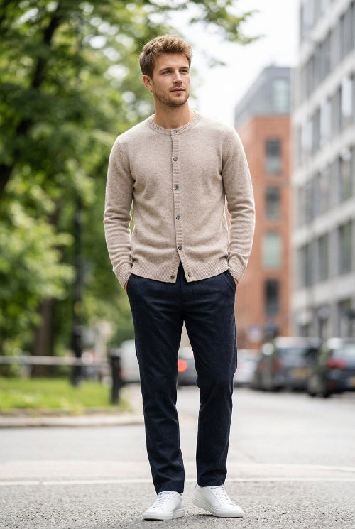 A light beige knit cardigan sweater with a crew neck and button closure, hanging on a dark wooden hanger against a solid gray background.