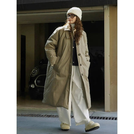Woman wearing an open army green long padded coat with her hands in the side pockets, styled with white wide-leg trousers and a knit hat