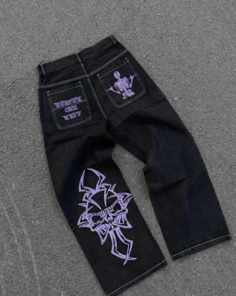 Back view of black wide-leg jeans laid flat, featuring purple lettering and a skeleton graphic on the back pockets and a large purple cross-style design on one leg.