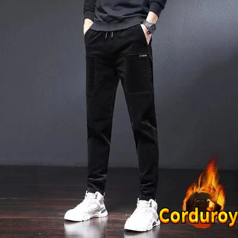 A person wearing black corduroy pants with large patch pockets, a dark sweatshirt, and white sneakers, standing against a dark gray wall.