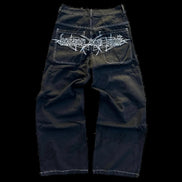 Back view of black wide-leg jeans laid flat, featuring white contrast stitching and a large white gothic lettering graphic across both back pockets.