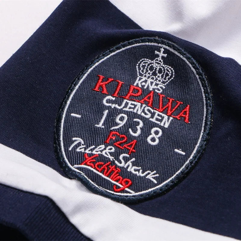 Close-up of a navy blue embroidered patch on a white and navy striped garment. The patch features a crown, the text 'KINS KI PAWA CJJENSEN 1938 F24 Talk & Shark Kicking' in red and white embroidery.