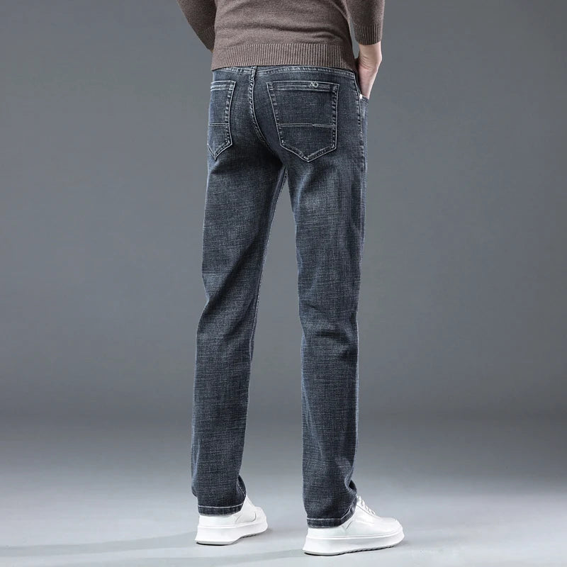 A man shown from the back wearing dark gray slim-fit jeans, a brown ribbed sweater, and white sneakers against a solid gray studio background.