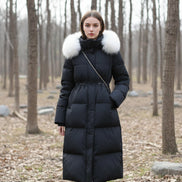 A person takes a mirror selfie wearing a long black puffer coat with a large light beige fur-trimmed collar. 
