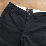 Black chino pants laid flat on a light brown wooden surface, showing the front waistband, button closure, belt loops, and side pockets.