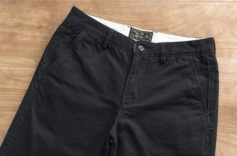 Black chino pants laid flat on a light brown wooden surface, showing the front waistband, button closure, belt loops, and side pockets.