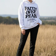 White hoodie with 'Faith Over Fear' text on a concrete surface with sneakers and a plant.