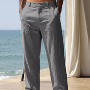 A man wearing loose, light gray linen trousers stands barefoot on a patio with the ocean and a cream curtain in the background.