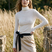 A person wearing a fitted long-sleeve white mock-neck or turtleneck top with a black belt at the waist, shown in a mirror selfie-style indoor photo.