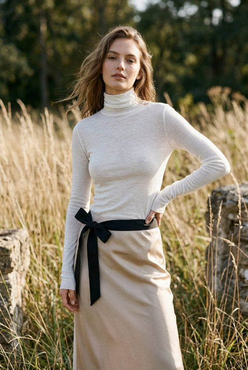 A person wearing a fitted long-sleeve white mock-neck or turtleneck top with a black belt at the waist, shown in a mirror selfie-style indoor photo.
