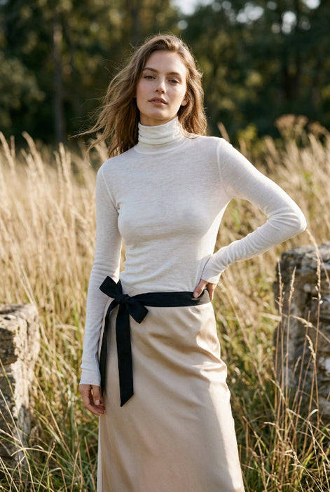 A person wearing a fitted long-sleeve white mock-neck or turtleneck top with a black belt at the waist, shown in a mirror selfie-style indoor photo.