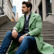 A man wearing a light green double-breasted overcoat, dark gray button-up shirt, white t-shirt, and blue jeans sits on outdoor concrete stairs in an urban setting.