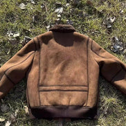 A brown suede shearling jacket with a dark brown ribbed knit collar, cuffs, and hem, laid flat on a grassy ground scattered with dry leaves.