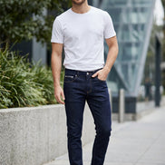 Dark blue slim-fit jeans hanging on a wooden hanger, with a white t-shirt partially visible behind them against a dark gray wall.