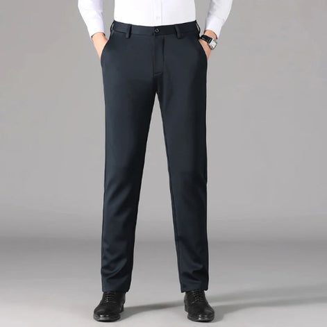 A man wearing slim-fit dark navy trousers, a white dress shirt, and black dress shoes, standing against a neutral gray studio background.