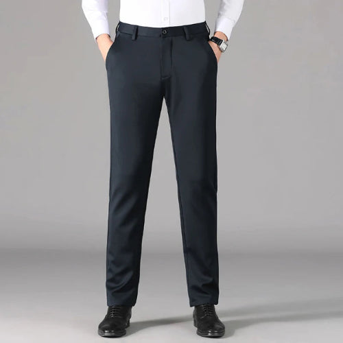 A man wearing slim-fit dark navy trousers, a white dress shirt, and black dress shoes, standing against a neutral gray studio background.