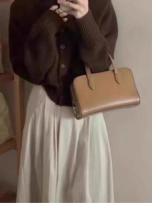A person taking a mirror selfie wearing a dark brown ribbed knit cardigan, a long cream-colored pleated skirt, and carrying a structured tan leather handbag.