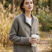 A woman wearing a cozy gray knit cardigan over a white top, holding a white paper coffee cup in a softly lit indoor setting.