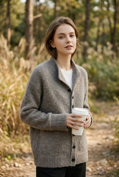 A woman wearing a cozy gray knit cardigan over a white top, holding a white paper coffee cup in a softly lit indoor setting.