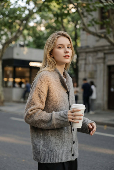 A woman wearing a cozy gray knit cardigan over a white top, holding a white paper coffee cup in a softly lit indoor setting.