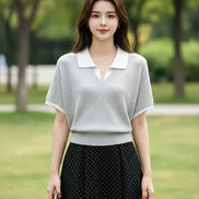 A woman wearing a light gray ribbed short-sleeve polo knit top with a white collar, holding a smartphone and carrying a black shoulder bag.