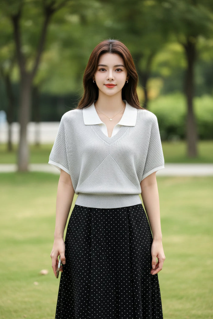 A woman wearing a light gray ribbed short-sleeve polo knit top with a white collar, holding a smartphone and carrying a black shoulder bag.