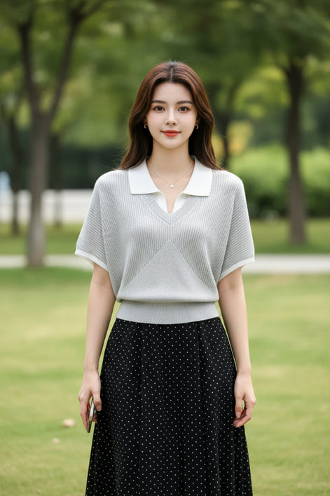 A woman wearing a light gray ribbed short-sleeve polo knit top with a white collar, holding a smartphone and carrying a black shoulder bag.