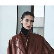 Close-up of a woman wearing a reddish-brown faux leather jacket with layered collars over a black turtleneck beside a window