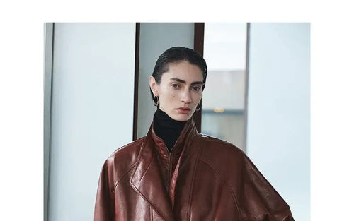Close-up of a woman wearing a reddish-brown faux leather jacket with layered collars over a black turtleneck beside a window