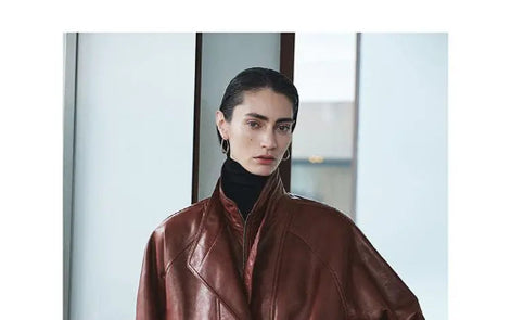 Close-up of a woman wearing a reddish-brown faux leather jacket with layered collars over a black turtleneck beside a window