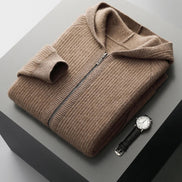 A folded taupe-brown ribbed knit zip-up sweater with a hood, displayed on a dark gray pedestal alongside a black leather strap watch.